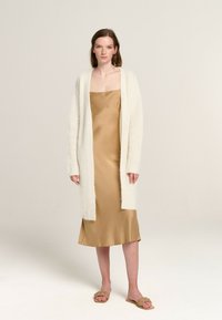Cremfarbener Cardigan mit offenem Vorderteil über einem goldenen Satin- Slipdress, kombiniert mit beigen, geflochtenen Slidesandalen. Sanfte Texturen und minimalistisches Design.