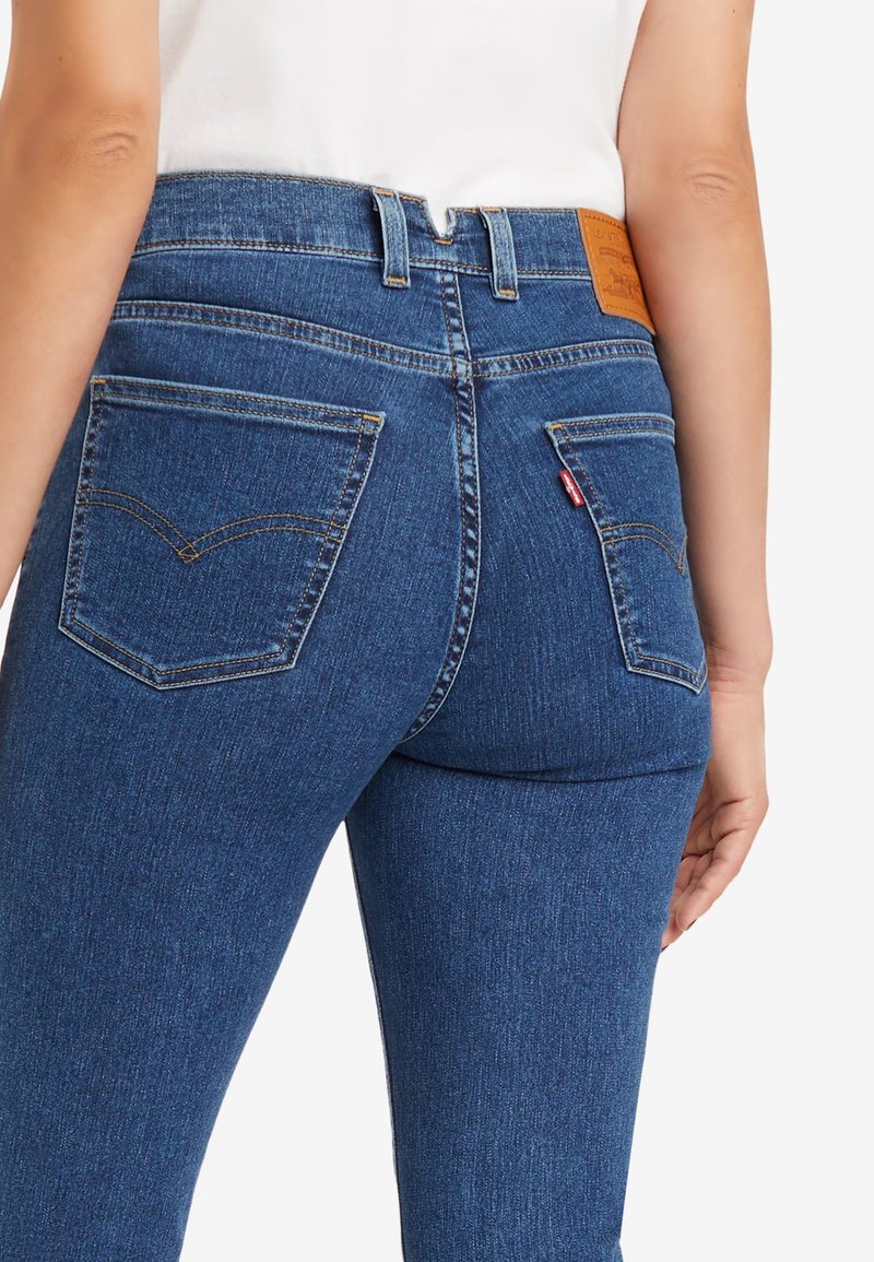 Jeans en denim bleu taille haute avec une texture lisse, dotés de deux poches arrière, de coutures jaunes et d'une étiquette en cuir de marque au niveau de la taille.
