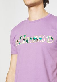 Fialové bavlněné tričko s různobarevným logem "ellesse" a různými vzory. Krátké rukávy a kulatý výstřih. Hladká textura.