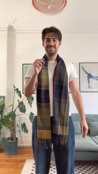 Écharpe à motifs tartans en vert, navy et gris avec des bords à franges, portée sur un t-shirt blanc. L'arrière-plan présente une plante verte et un canapé clair.