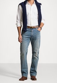 Jeans in denim azzurro, camicia bianca con bottoni, maglione in caldo cotone blu navy drappeggiato sulle spalle, cintura in pelle marrone e stivali marroni.