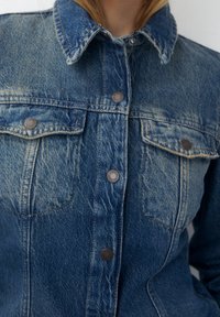 Chaqueta de mezclilla en azul claro con una textura deslavada, que presenta un collar clásico, bolsillos frontales y botones de metal marrón.