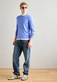 Hellblaue langärmlige Pullover mit gerippten Bündchen, über einem weißen T-Shirt getragen. Blue Jeans und grün gestreifte Turnschuhe vervollständigen das Outfit.