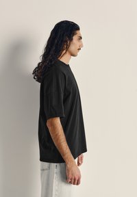 Černé bavlněné tričko s krátkým rukávem, uvolněného střihu a kulatým výstřihem. Model nosí světlé džínové kalhoty.