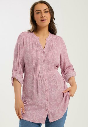 Donna che indossa una camicetta rosa chiaro a maniche lunghe con bottoni e motivi floreali bianchi, abbinata a jeans blu, in piedi davanti a uno sfondo semplice.