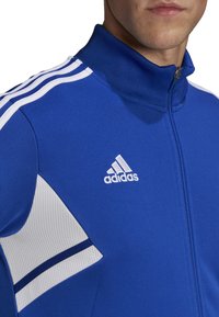 Blue zip-up jacket made of textured fabric. Features white stripes on shoulders and an embroidered Adidas logo on the chest.