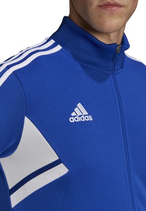 Blauwe ritsjacket gemaakt van textuurstof. Heeft witte strepen op de schouders en een geborduurd Adidas-logo op de borst.