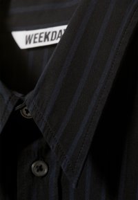 Musta paita, jossa on tummansiniset pystysuuntaiset raidat, napitettava etuosa, terävä kaulus ja kankainen etiketti, jossa lukee "WEEKDAY" kaula-aukon sisäpuolella.