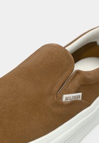 Καφέ παπούτσι από σουέντ με λευκή σόλα και μικρή υφασμάτινη ετικέτα με την ένδειξη «Hilfiger» κοντά στο ελαστικό πάνελ στο πλάι.
