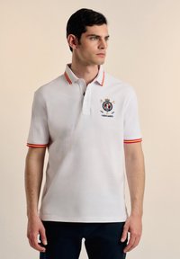 Camisa polo blanca con cuello y puños de mangas a rayas rojas y amarillas. Presenta un logo bordado en el pecho, mangas cortas y un corte clásico.