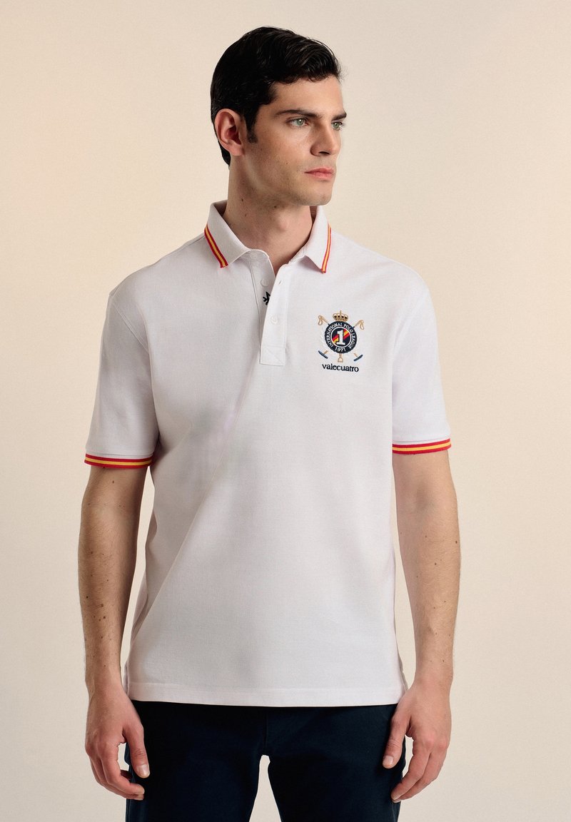 Camisa polo blanca con cuello y puños de mangas a rayas rojas y amarillas. Presenta un logo bordado en el pecho, mangas cortas y un corte clásico.