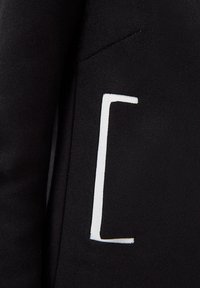 Chaqueta de tela negra con un prominente detalle de logo blanco, líneas elegantes y textura suave. No se observan adornos adicionales.
