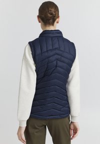 Mornarsko puffer telovnik z vzorcem chevron, visokim ovratnikom in armhole-om. Uporabljen čez svetlo sivo pulover in olivno zelene hlače.