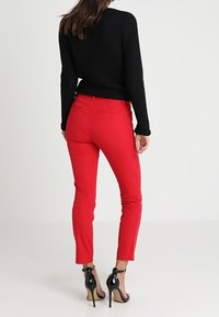 Vrouw in een zwart gebreid sweater, rode slim-fit broek en zwarte hoge hakken, staande met de rug naar de camera op een effen witte achtergrond.