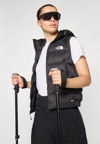 Zwarte puffy vest met rits, logo op de borst, gedragen over een wit shirt. Zwarte cargobroek en trekkingstokken, geaccessoireerd met grote zonnebril.