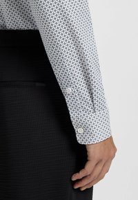 White shirt with a navy geometric pattern, long sleeves, button cuffs, and a textured black fabric in the background.