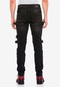 Schwarze Denim-Jeans mit schmaler Passform, die ein teilweise zerrissenes Design, mehrere Taschen und Gürtelschlaufen aufweisen. Rote und schwarze Stiefel sind ebenfalls sichtbar.
