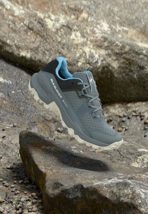 Harmaa ja musta Mammutin polkujuoksukenkä vaaleansinisen vuorauksen kanssa, leijailemassa kivisen ulkoilmamaaston yläpuolella.