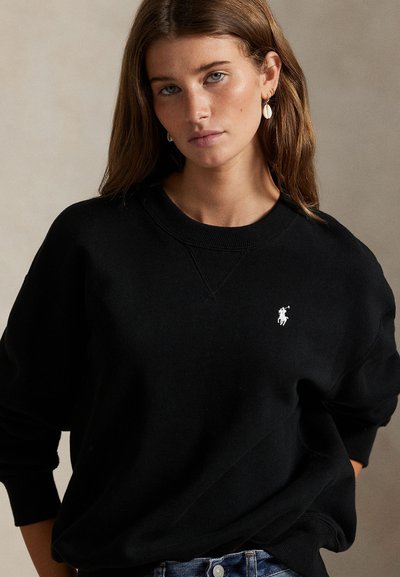 Ung kvinne med lys brunt hår som har på seg en svart genser med en liten hvit hestelogo og skjelløreringer mot beige bakgrunn.