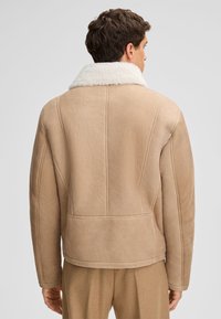 Giacca marrone in shearling con colletto in eco pelliccia bianca, caratterizzata da una forma squadrata, superficie testurizzata e dettagli di cuciture visibili sulla schiena.