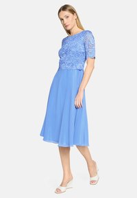 Robe bleue avec un haut en dentelle, manches courtes et une jupe fluide. Le tissu a une texture lisse, et le design est longueur genou.