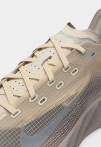 Κοντινό πλάνο μπεζ αθλητικού παπουτσιού Nike με σχέδιο πλέγματος, ανοιχτό μπεζ κορδόνια και διαφανές ανώτερο υλικό.