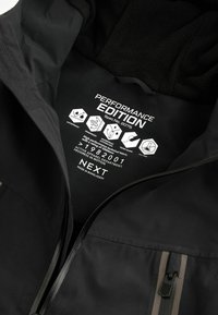 Veste imperméable noire dotée d'un intérieur en polaire, de poches zippées et d'étiquettes d'édition performance avec instructions d'entretien.
