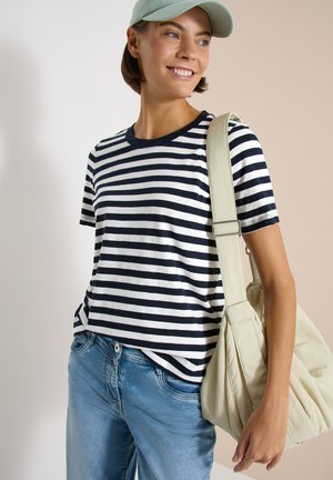 Kvinde smilende iført en mintgrøn kasket, sort-hvidstribet t-shirt, blå jeans, bærende en stor beige skuldertaske.