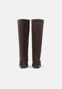 Bottes en suède marron montantes jusqu'au genou, avec une texture lisse, des talons carrés et un minimum de détails de couture. Design simple et élégant sans embellissements.
