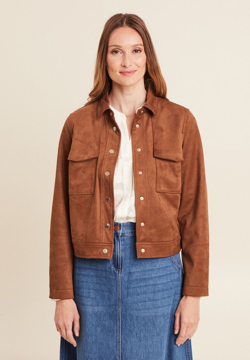 Veste en daim marron avec un devant boutonné, deux poches poitrine et une coupe courte. Portée sur une chemise légère avec une jupe en denim.
