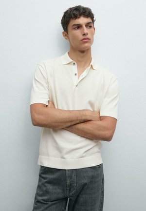 Young man with folded arms wearing a cream short-sleeve polo shirt and dark jeans against a plain light gray background.