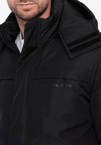 Černá zateplená bunda s vysokým límcem, zipovým uzávěrem, texturovanou tkaninou a suchými zipy na ramenou. Disponuje decentním logem.