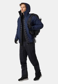 Marinblå vattentät jacka med justerbar huva, över en svart isolerad väst, tillsammans med mörka byxor och svarta handskar. Utrustad med en ryggsäck.