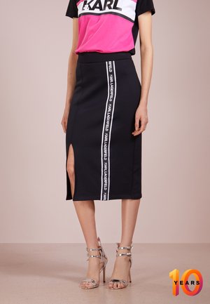 Kvinde iført en sort blyantsnederdel med en vertikal Karl Lagerfeld-logo stribe og sideslidser, kombineret med sølvfarvede høje hæle sandaler.