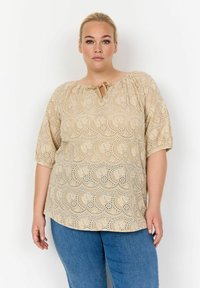 Top beige con scollatura tonda e dettaglio a chiave, caratterizzato da un motivo floreale a taglio e maniche corte a palloncino, abbinato a jeans blu.