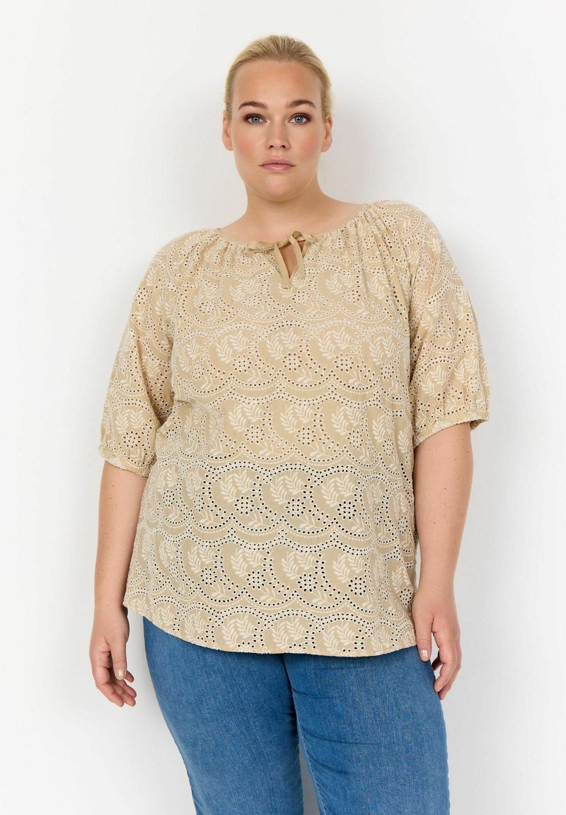 Top beige con scollatura tonda e dettaglio a chiave, caratterizzato da un motivo floreale a taglio e maniche corte a palloncino, abbinato a jeans blu.
