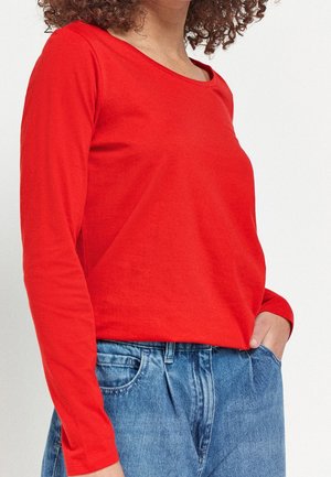 Osoba ubrana w jasnoczerwoną bluzkę z długim rękawem, wpuszczoną do wysokich dżinsów z niebieskiego denimu, z ręką włożoną do kieszeni na jednolitym tle.