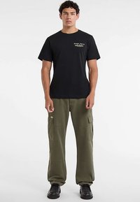 T-shirt noir à col ras du cou avec un logo sur le côté gauche de la poitrine, associé à un pantalon cargo olive avec plusieurs poches et une coupe décontractée.