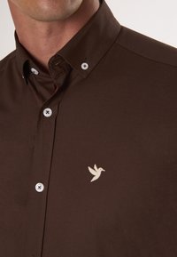 Camicia marrone con bottoni, caratterizzata da un colletto, bottoni bianchi e un piccolo logo a forma di uccello color crema ricamato sul lato sinistro.