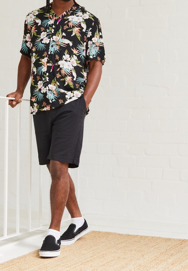 Camicetta nera a maniche corte con fiori colorati, abbinata a pantaloni corti scuri e scarpe slip-on nere con suole bianche.