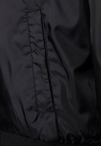 Černá bomber bunda s detaily, obsahující boční kapsu, hladkou nylonovou texturu a žebrovaný lem. Šité švy jsou viditelné po celé tkanině.