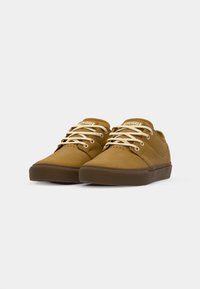 Brown canvas sneakers with cream laces, low profile, rounded toe, and textured rubber sole. Brand name visible on the tongue.
