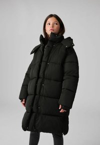 Melnā pūkaina jaka ar quilted dizainu, augstu apkakli un ar rāvējslēdzēju aizveramu priekšu. Jakai ir lielas, mīkstas daļas siltumizolācijai.