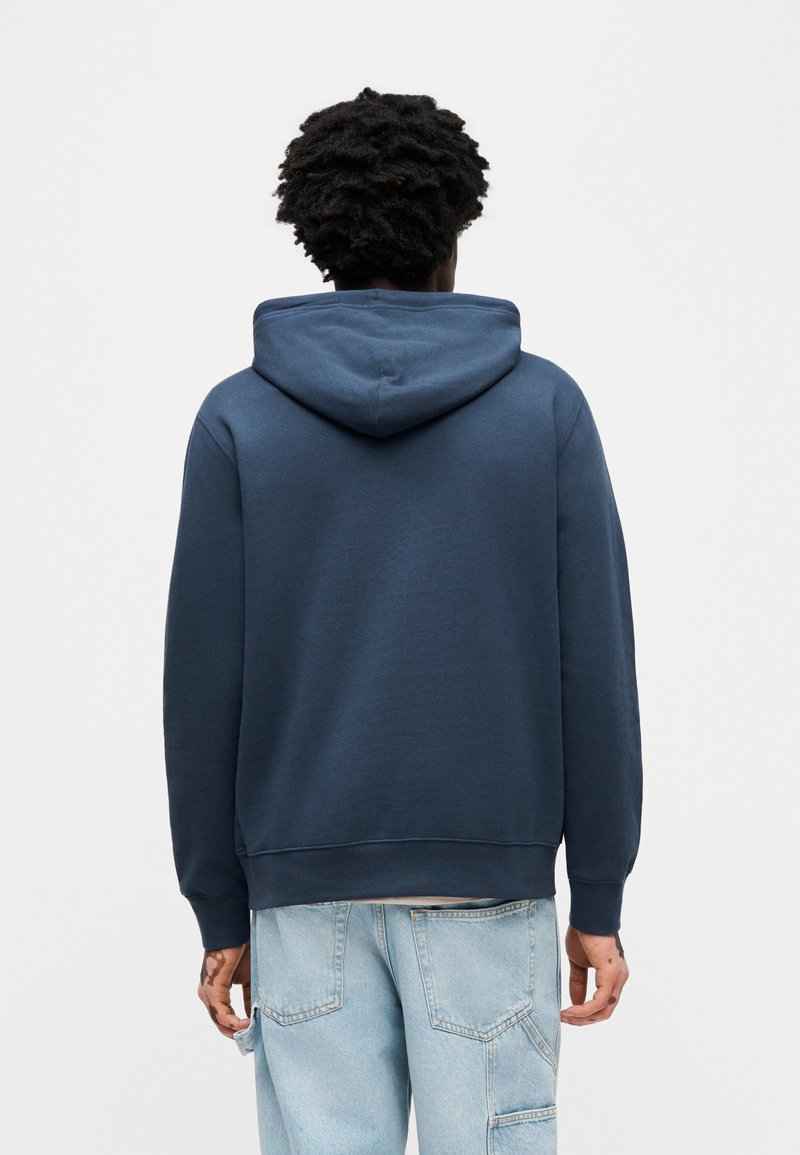 Marineblå hættetrøje med en rummelig hætte, ribstrikkede manchetter og kant. Har en glat tekstur og afslappet pasform, vist fra bagsiden.