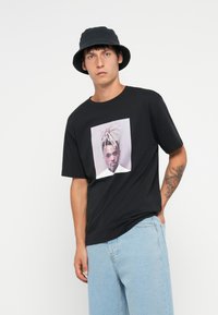 Svart bomull T-skjorte med et sentralt grafisk motiv av en person med lyst hår. Kombinert med lys blå denimshorts og en svart bøttehatt.