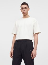 Jeune homme debout face à l'objectif, portant une chemise blanche à manches courtes unie et un pantalon noir avec un petit logo de couteau et fourchette croisés.