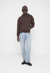 Καφέ φούτερ με κουκούλα και μπροστινή τσέπη, ανοιχτό μπλε τζιν σε ίσια γραμμή και μαύρα παπούτσια slip-on. Φορά επίσης ριγέ πλεκτό σκουφάκι.