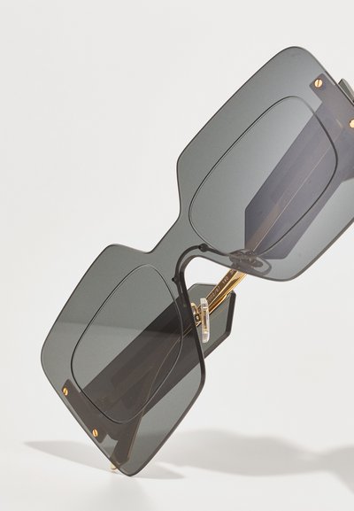 Lunettes de soleil surdimensionnées de forme carrée avec des verres gris foncé, monture noire et accents dorés. Elles présentent un design géométrique et du matériel en métal visible.