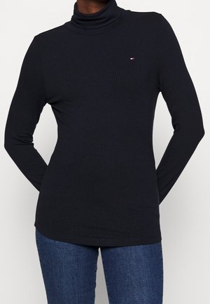 Personne portant un pull à col roulé côtelé noir ajusté avec un petit logo sur la poitrine et un jean en denim bleu.