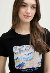 Zwarte t-shirt met een kleurrijke print van strandparasols en ligstoelen in blauwe, witte en zandtinten. De tekst luidt "italian vacay club."